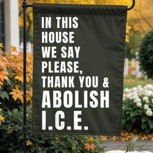 May include: A black garden flag with white text that reads "IN THIS HOUSE WE SAY PLEASE, THANK YOU & ABOLISH I.C.E." The flag is hanging on a black metal pole in front of a fall-themed background with pumpkins.