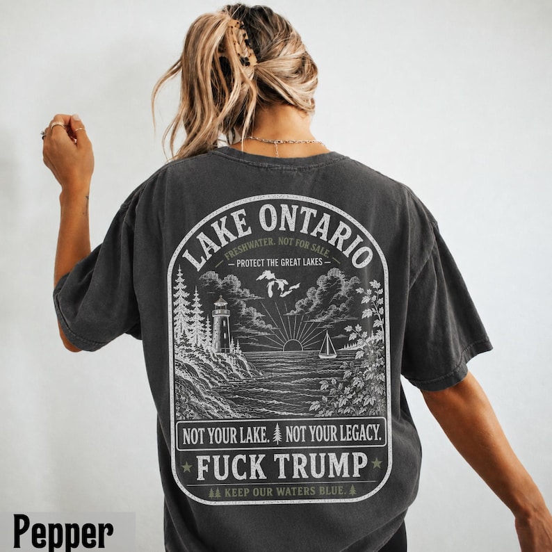 May include: A charcoal gray t-shirt featuring a white graphic on the back. The design depicts a lighthouse, a sailboat, and a map of the Great Lakes with the text: LAKE ONTARIO, FRESHWATER. NOT FOR SALE. PROTECT THE GREAT LAKES. NOT YOUR LAKE. NOT YOUR LEGACY. FUCK TRUMP. KEEP OUR WATERS BLUE.