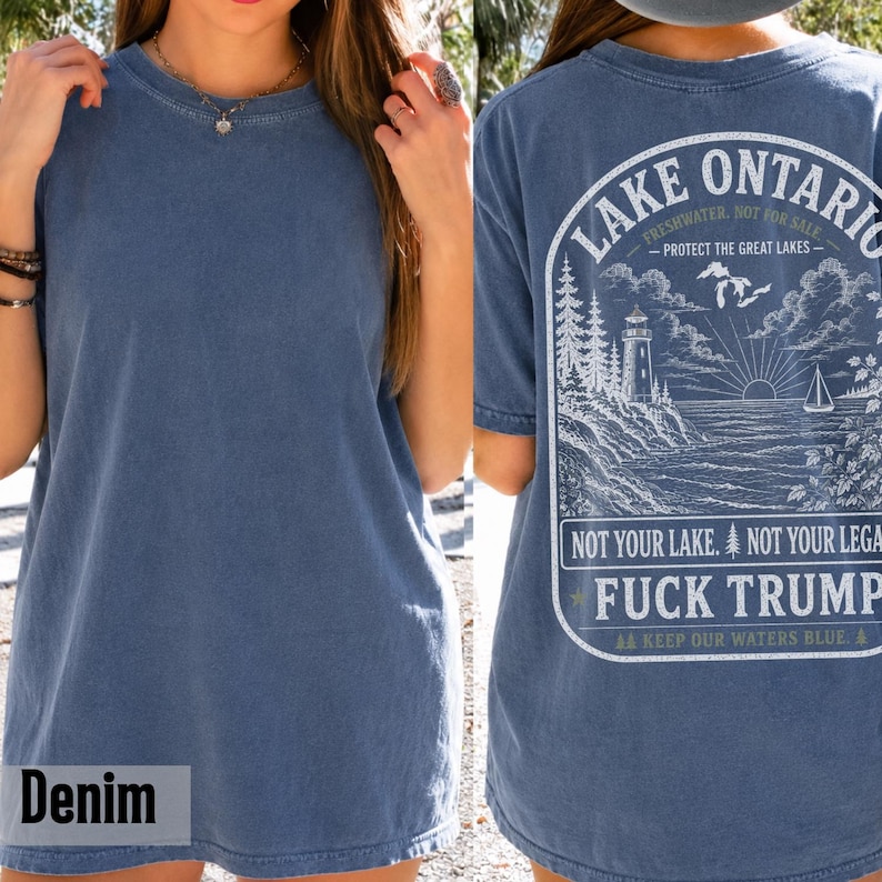May include: A denim blue t-shirt featuring a white graphic on the back. The design depicts a lighthouse, a sailboat, and a sunburst over water with the text Lake Ontario, Freshwater, Not For Sale, Protect The Great Lakes, Not Your Lake, Not Your Legacy, Fuck Trump, and Keep Our Waters Blue.