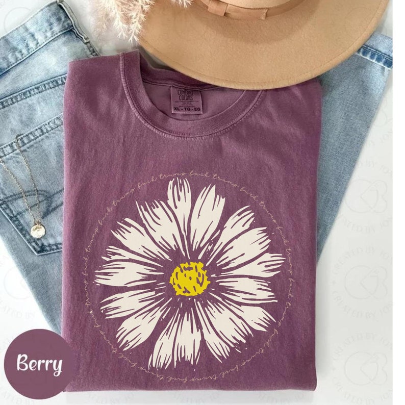 May include: A purple t-shirt with a white daisy graphic and the text "back, trust, back, trust, back, trust, back, trust" repeated around the daisy. The shirt is folded and laid on a pair of blue jeans.