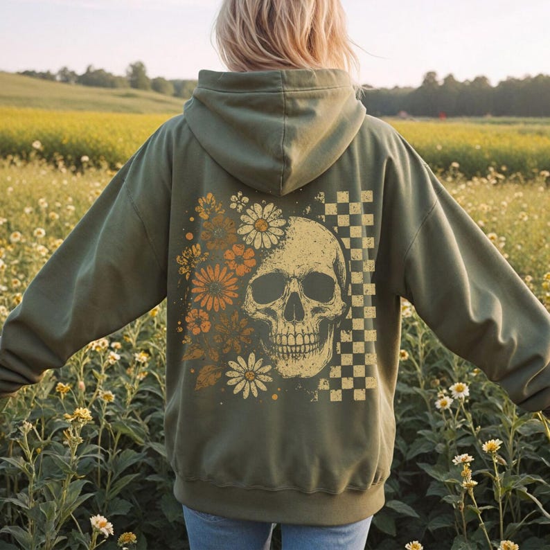 May include: Olive green hoodie with a skull graphic, flowers, and a checkered pattern. The back of the hoodie features a detailed illustration of a skull with floral elements in shades of orange, white, and yellow. The design is on the back of the hoodie.