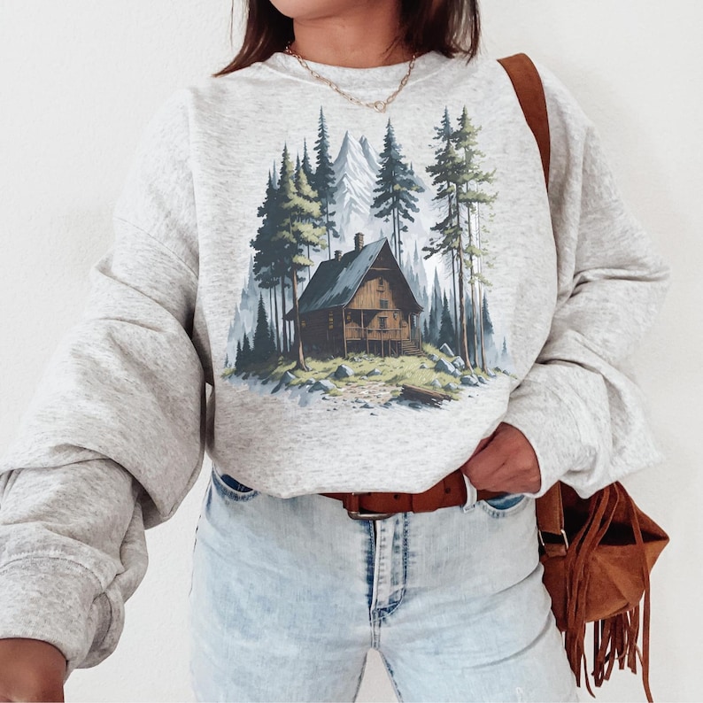 May include: A light gray sweatshirt featuring a cabin scene in a forest with tall green trees and a mountain backdrop. The cabin is brown with a dark blue roof. The sweatshirt has long sleeves and a crew neck.