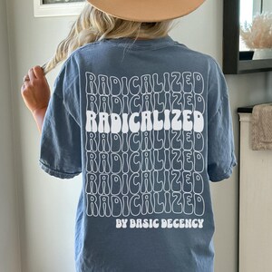 May include: A blue t-shirt with the word "RADICALIZED" stacked multiple times in a white, retro font on the back. Below the text is "BY BASIC DECENCY" in a smaller font. The shirt is a solid color and has short sleeves.