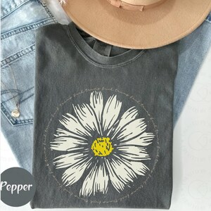 May include: A dark gray t-shirt with a white daisy graphic and the text "find your own truth, find your own light, find your own truth, find your own light" in a circle around the daisy.