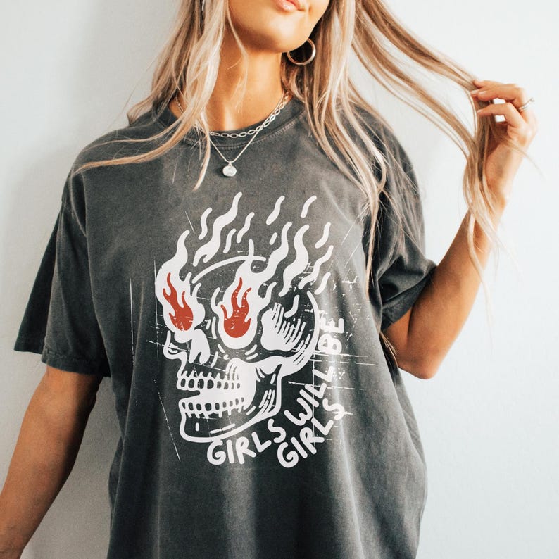 May include: A dark grey t-shirt with a skull graphic and the text "Girls Will Be Girls" printed on the front. The skull has flames coming out of its eyes.