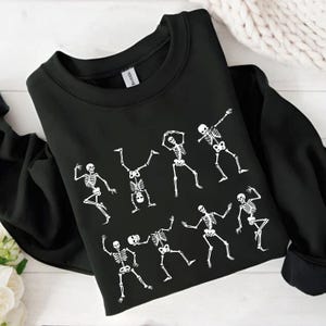 K&ouml;nnte beinhalten: Schwarzes Sweatshirt mit Rundhalsausschnitt und wei&szlig;em Skelett-Design. Das Design zeigt mehrere Skelette in verschiedenen Tanzposen. Das Sweatshirt ist aus weichem Material und ideal f&uuml;r Halloween.
