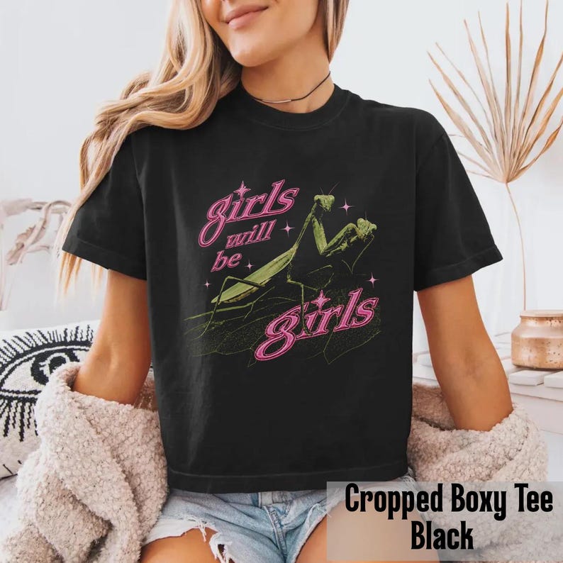 以下が含まれることがあります： 緑色のカマキリのグラフィックが描かれた黒の半袖Tシャツ。昆虫の上下にはピンクのネオン風フォントで「girls will be girls」という文字がプリントされており、デザイン全体に小さなピンクの星の形がアクセントとして散りばめられています。