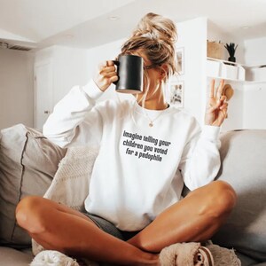 May include: White sweatshirt with long sleeves and the text "imagine telling your children you voted for a pedophile" in black. A black mug is held up to the face. The person is sitting on a couch.
