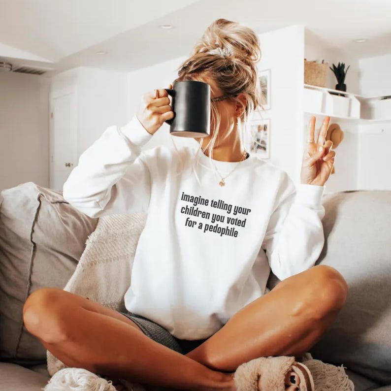 May include: White sweatshirt with long sleeves and the text "imagine telling your children you voted for a pedophile" in black. A black mug is held up to the face. The person is sitting on a couch.