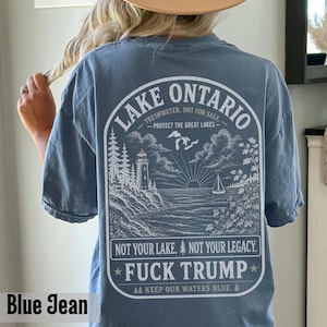 May include: A blue jean colored t-shirt featuring a white graphic on the back. The design depicts a lighthouse, trees, a sailboat, and the text: LAKE ONTARIO FRESHWATER. NOT FOR SALE. PROTECT THE GREAT LAKES. NOT YOUR LAKE. NOT YOUR LEGACY. FUCK TRUMP.