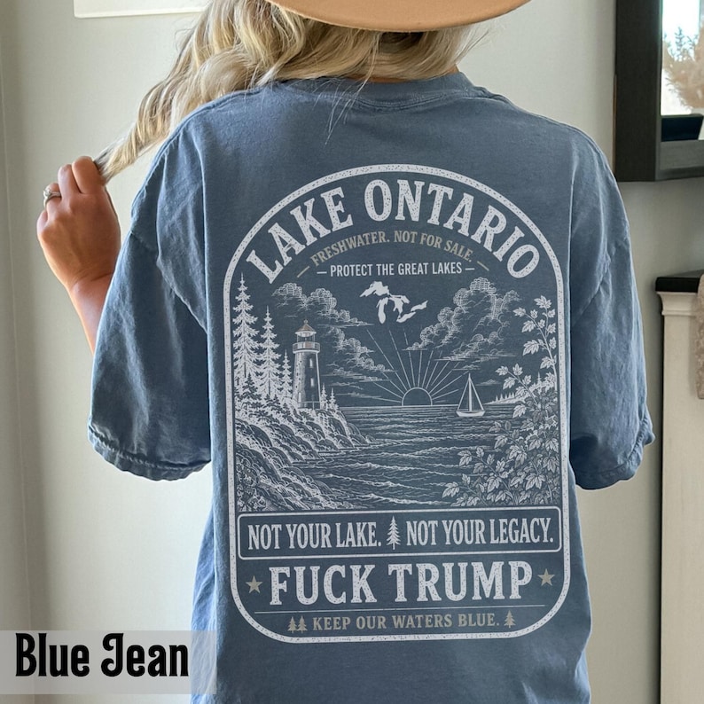 May include: A blue jean colored t-shirt featuring a white graphic on the back. The design depicts a lighthouse, trees, a sailboat, and the text: LAKE ONTARIO FRESHWATER. NOT FOR SALE. PROTECT THE GREAT LAKES. NOT YOUR LAKE. NOT YOUR LEGACY. FUCK TRUMP.