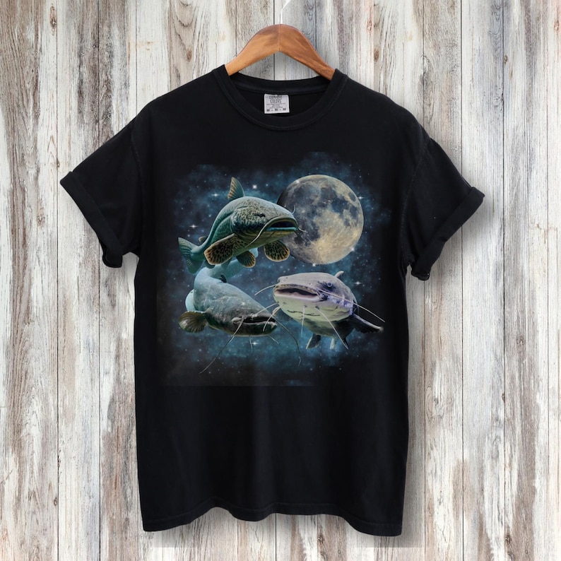 May include: Black t-shirt with a graphic print of three catfish swimming in a starry night sky with a full moon.