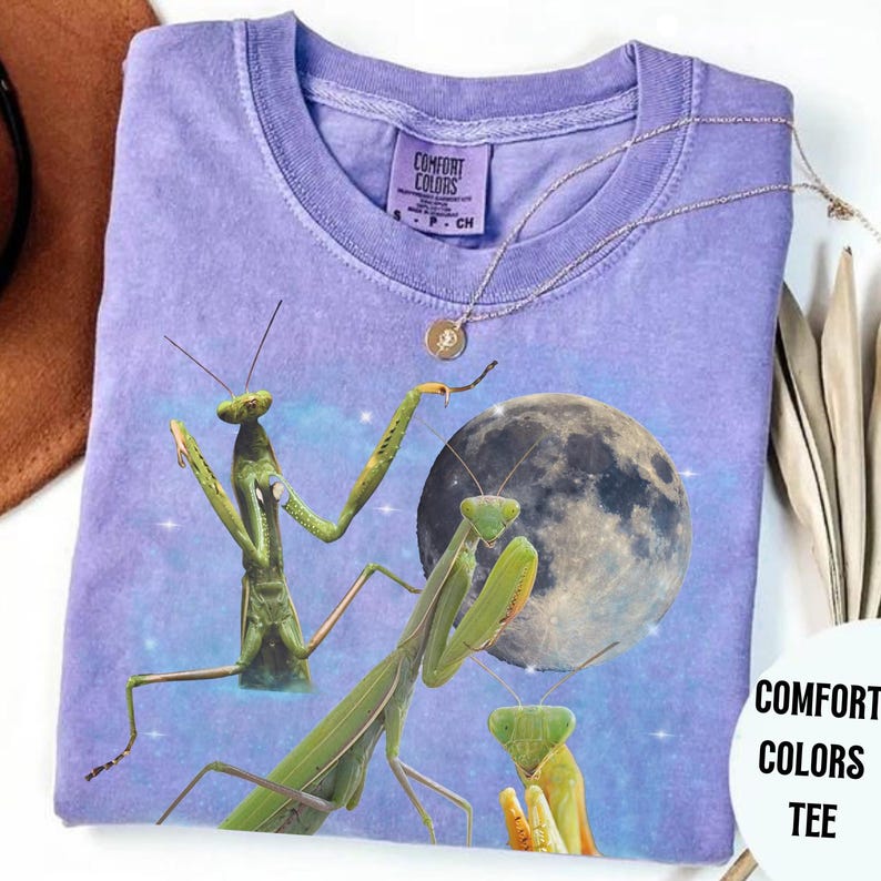 Puede incluir: Una camiseta Comfort Colors lavanda con un gr&aacute;fico de tres mantis religiosas verdes y una luna llena. La camiseta tiene cuello redondo y mangas cortas. Un collar dorado est&aacute; drapeado sobre la camiseta.