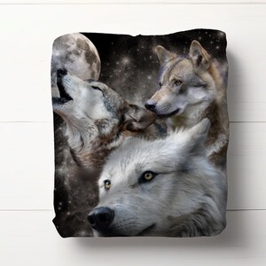 May include: A plush blanket featuring a nighttime scene with three wolves. One wolf is howling at a full moon, while the others look on. The background is a starry night sky with a dark color palette.