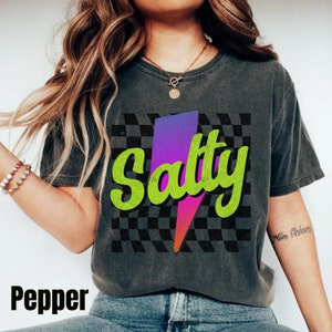 以下が含まれることがあります： チェック柄の背景に、稲妻のグラフィックと緑色の文字で「Salty」と書かれた黒いTシャツです。