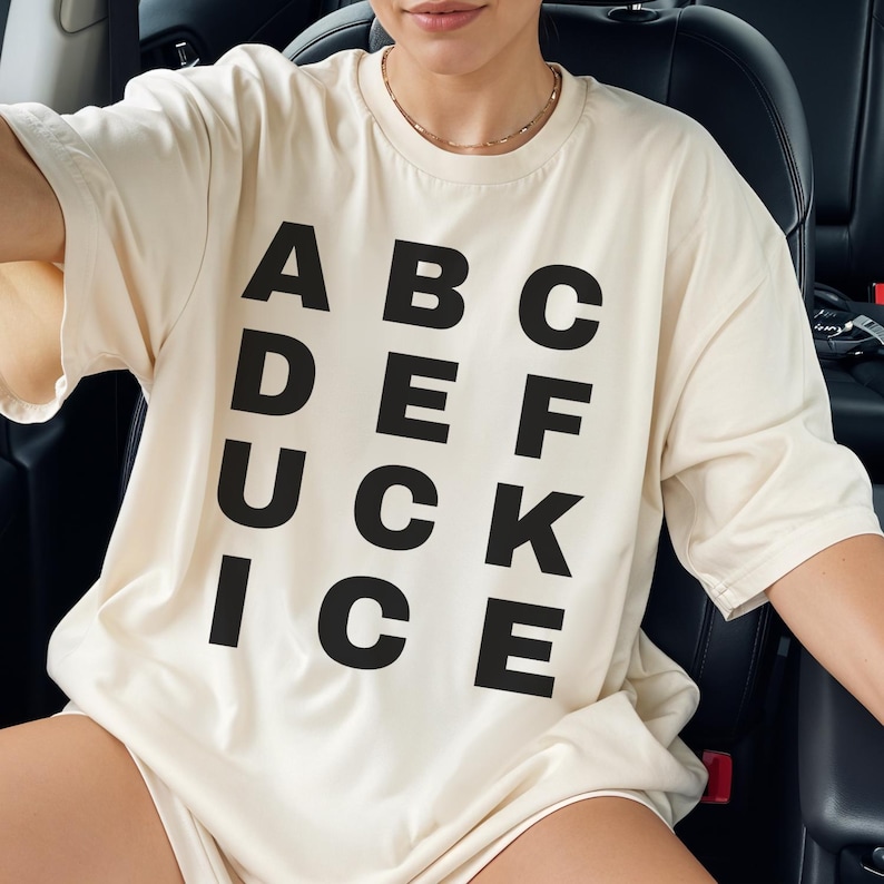 May include: Cream-colored t-shirt with the word "ADUCKICE" stacked vertically on the left side and "ABC", "DEF", and "CKE" stacked vertically on the right side in black block letters. The shirt is oversized.