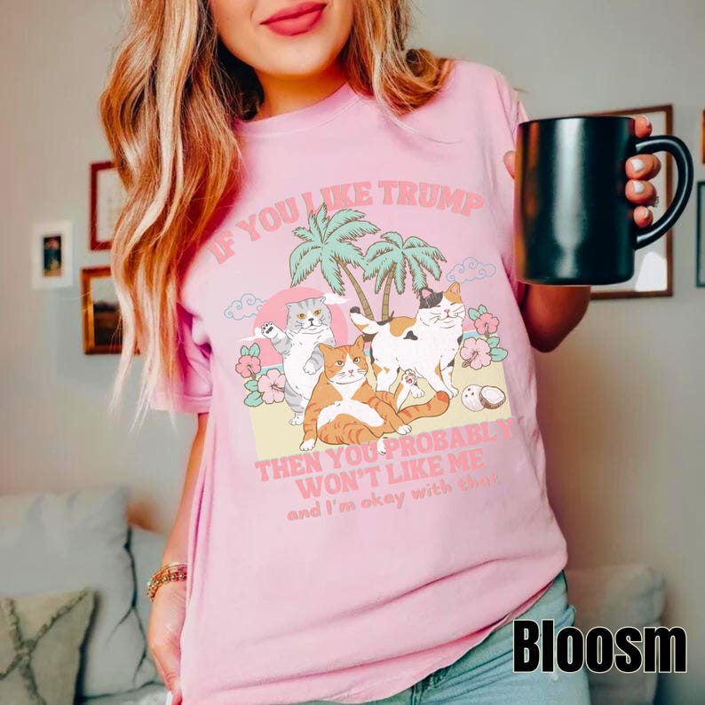 May include: Pink t-shirt with a graphic of cats on a beach with palm trees and the text "If you like Trump then you probably won't like me and I'm okay with that."