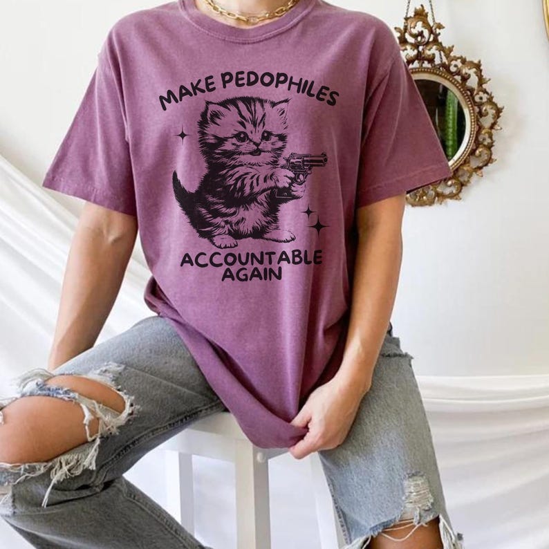 Op de afbeelding: Een mauve t-shirt met de tekst "MAKE PEDOPHILES ACCOUNTABLE AGAIN" en een afbeelding van een katje met een pistool. Het shirt wordt gedragen met een gescheurde blauwe jeans.
