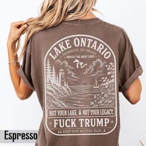May include: An espresso-colored t-shirt featuring a white graphic on the back. The design depicts a lighthouse, a sailboat, and a sunburst over Lake Ontario. Text includes "LAKE ONTARIO," "PROTECT THE GREAT LAKES," "NOT YOUR LAKE. NOT YOUR LEGACY. FUCK TRUMP," and "KEEP OUR WATERS BLUE."