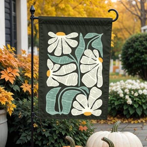 May include: A green garden flag featuring a white floral design. The flowers are stylised and have a textured appearance. The flag is hanging from a black metal pole.