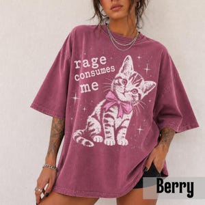 May include: A berry-colored oversized t-shirt featuring a white graphic of a kitten wearing a pink bow. The text "rage consumes me" is printed in white above the kitten, surrounded by small star accents. The shirt has short sleeves and a crew neck.