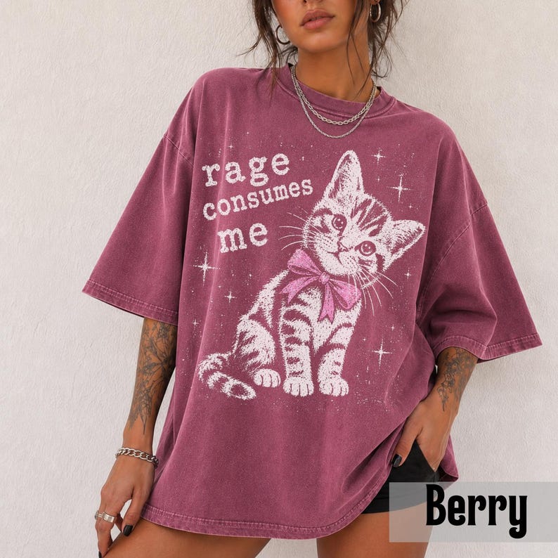 May include: A berry-colored oversized t-shirt featuring a white graphic of a kitten wearing a pink bow. The text "rage consumes me" is printed in white above the kitten, surrounded by small star accents. The shirt has short sleeves and a crew neck.
