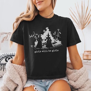 Pu&ograve; includere: Una t-shirt nera a maniche corte con una stampa grafica bianca di figure con cappelli a punta attorno a un fal&ograve;. Il testo "girls will be girls" &egrave; stampato con un carattere simile a quello di una macchina da scrivere sotto l'immagine.