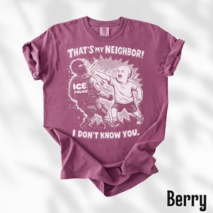K&ouml;nnte beinhalten: Beerenfarbenes T-Shirt mit einer grafischen Illustration. Die Illustration zeigt ein Kind, das einen ICE-Polizisten schl&auml;gt, mit dem Text "THAT'S MY NEIGHBOR! I DON'T KNOW YOU." ober- und unterhalb des Bildes.