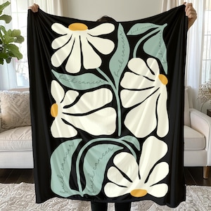 May include: Black blanket featuring a floral design with large white daisies, yellow centers, and light blue leaves. The words "fuck cancer" are written in a script font. The blanket is held up by a person.