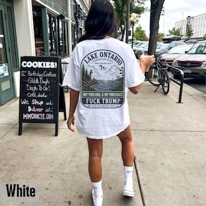 May include: A white t-shirt featuring a graphic on the back with a landscape illustration of Lake Ontario, a lighthouse, and a sailboat. The text reads Lake Ontario, Not Your Lake, Not Your Legacy, Fuck Trump, and Keep Our Waters Blue.