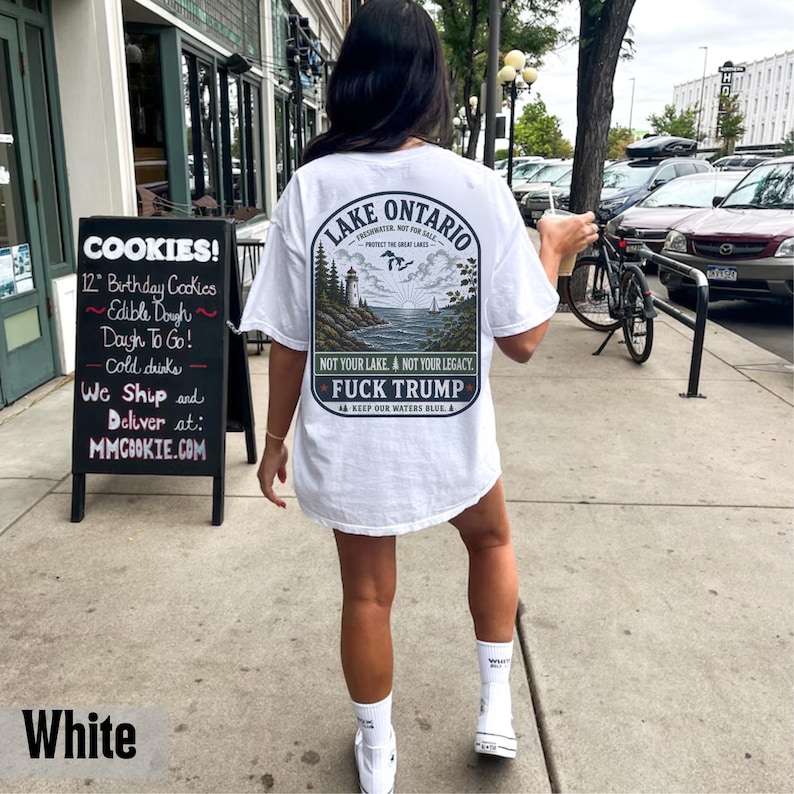 May include: A white t-shirt featuring a graphic on the back with a landscape illustration of Lake Ontario, a lighthouse, and a sailboat. The text reads Lake Ontario, Not Your Lake, Not Your Legacy, Fuck Trump, and Keep Our Waters Blue.