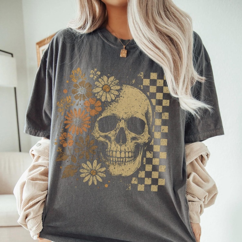 May include: Dark gray t-shirt featuring a skull graphic with floral and checkered design elements. The skull is in a vintage style, with orange and yellow flowers on the left side and a checkered pattern on the right.