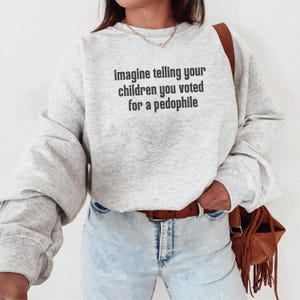 May include: A light gray sweatshirt with the text "imagine telling your children you voted for a pedophile" in black. The sweatshirt is paired with light wash jeans and a brown belt.