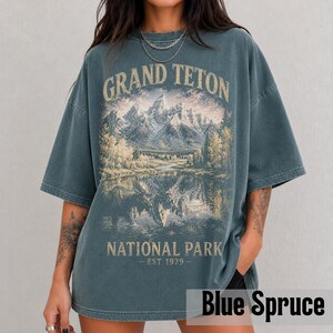 May include: Blue spruce-colored t-shirt featuring a scenic illustration of the Grand Teton National Park. The design includes mountains, a lake, and trees, with the text "GRAND TETON NATIONAL PARK - EST 1929." The shirt has a relaxed fit.