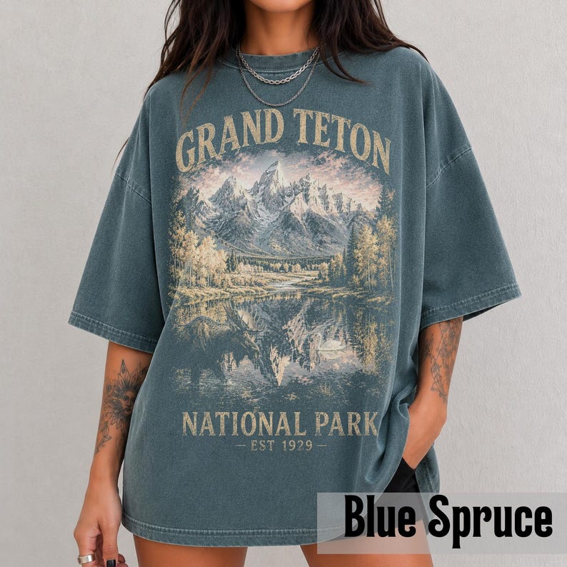 May include: Blue spruce-colored t-shirt featuring a scenic illustration of the Grand Teton National Park. The design includes mountains, a lake, and trees, with the text "GRAND TETON NATIONAL PARK - EST 1929." The shirt has a relaxed fit.