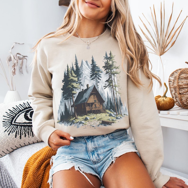 May include: Beige sweatshirt featuring a cabin scene in a forest with tall green trees and a mountain backdrop. The cabin is brown with a porch. The sweatshirt is worn with denim shorts.