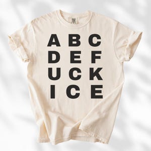 May include: Cream-colored t-shirt with the word "DUCK" spelled out vertically in bold, black capital letters, with other letters arranged around it. The shirt is made of a soft material and has rolled-up sleeves.