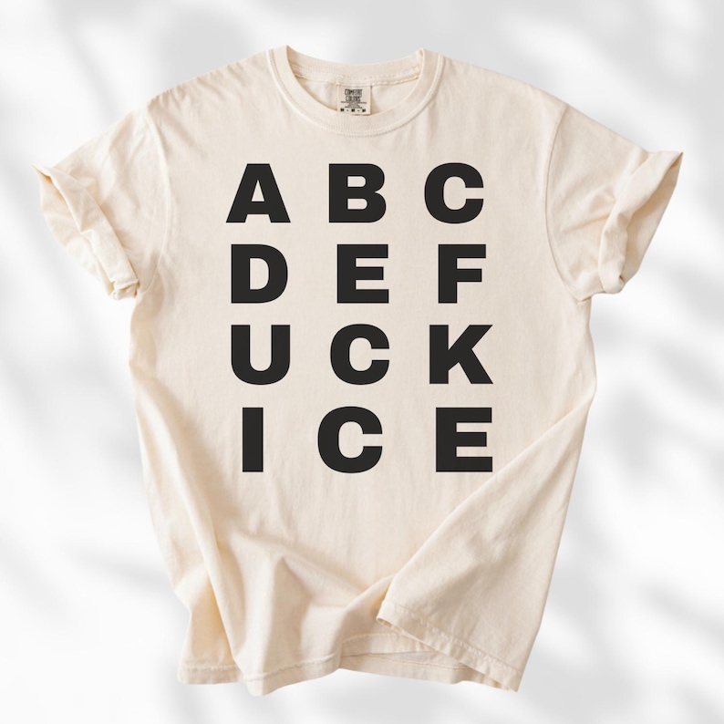 May include: Cream-colored t-shirt with the word "DUCK" spelled out vertically in bold, black capital letters, with other letters arranged around it. The shirt is made of a soft material and has rolled-up sleeves.