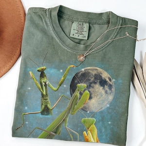 Puede incluir: Camiseta verde Comfort Colors con un gr&aacute;fico de tres mantis religiosas frente a una luna llena y un cielo estrellado. Un collar plateado con un peque&ntilde;o colgante descansa sobre la camiseta. La camiseta tiene una peque&ntilde;a etiqueta que dice "Comfort Colors".