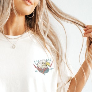 Op de afbeelding: Wit T-shirt met een afbeelding van een Amerikaanse zeearend in een driehoek met sterren en bliksemschichten. De adelaar heeft rode, blauwe en gele details.