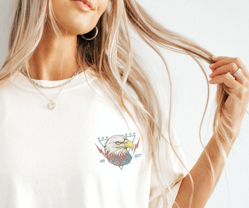 Op de afbeelding: Wit T-shirt met een afbeelding van een Amerikaanse zeearend in een driehoek met sterren en bliksemschichten. De adelaar heeft rode, blauwe en gele details.