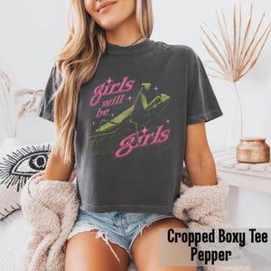 以下が含まれることがあります： チャコールグレーの半袖Tシャツ。緑色のカマキリのグラフィックと、小さな星のアクセントが付いたピンク色の「girls will be girls」という文字がプリントされています。ボックス型のクロップド丈で、クルーネックの柔らかい生地で作られています。