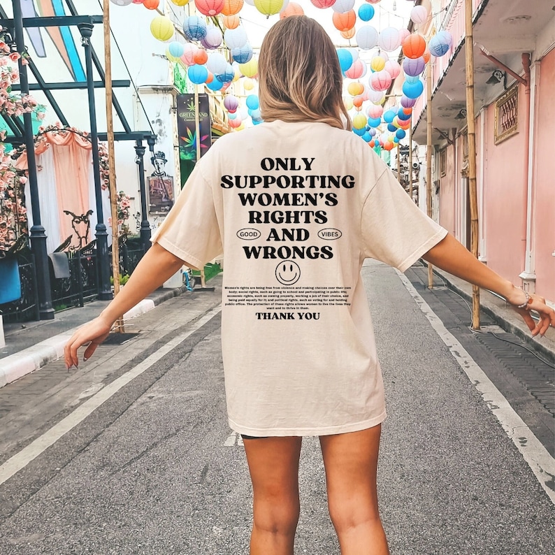 May include: A beige t-shirt with black text that reads "Only Supporting Women's Rights and Wrongs." The text is in a bold, sans-serif font. There is a smiley face below the text. The shirt is being worn by a person walking down a street.