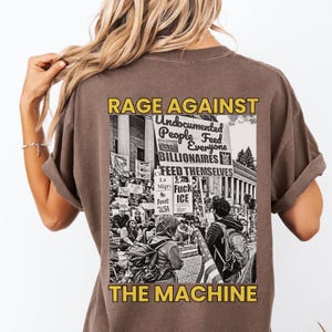 May include: Brown t-shirt with a graphic on the back. The graphic features the words "RAGE AGAINST THE MACHINE" in yellow, with a black and white image of a protest. The image includes text that reads "Undocumented People Feed Everyone Billionaires Feed Themselves."