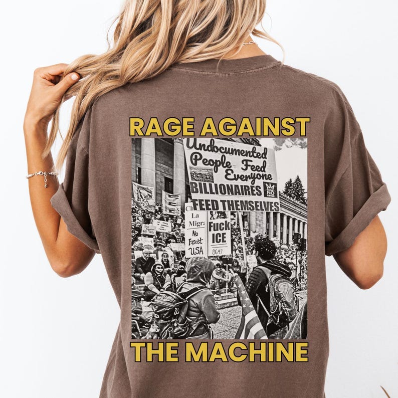 May include: Brown t-shirt with a graphic on the back. The graphic features the words "RAGE AGAINST THE MACHINE" in yellow, with a black and white image of a protest. The image includes text that reads "Undocumented People Feed Everyone Billionaires Feed Themselves."