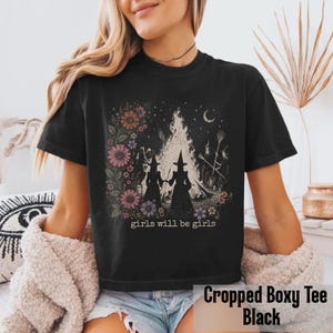May include: Black cropped boxy tee featuring a graphic of two witches in silhouette, a bonfire, and floral accents. The text "girls will be girls" is below the graphic. The shirt is a comfortable, casual style.