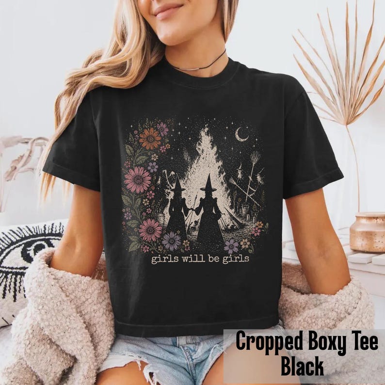 May include: Black cropped boxy tee featuring a graphic of two witches in silhouette, a bonfire, and floral accents. The text "girls will be girls" is below the graphic. The shirt is a comfortable, casual style.