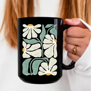 May include: Black ceramic mug with a white floral design. The flowers are stylized and have a yellow center. The leaves are green.