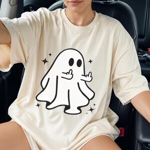 May include: Cream-colored t-shirt featuring a black and white cartoon ghost giving the middle finger. The ghost is surrounded by black star graphics. The shirt is a casual, oversized fit.