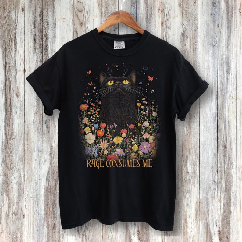 May include: Black t-shirt with a black cat in a field of colorful flowers. The text "RAGE CONSUMES ME" is printed below the cat.